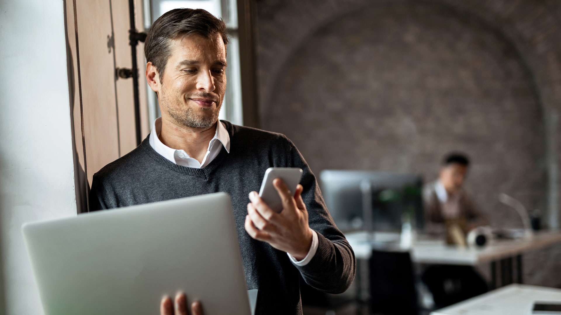 Business man looking at phone; holding laptop